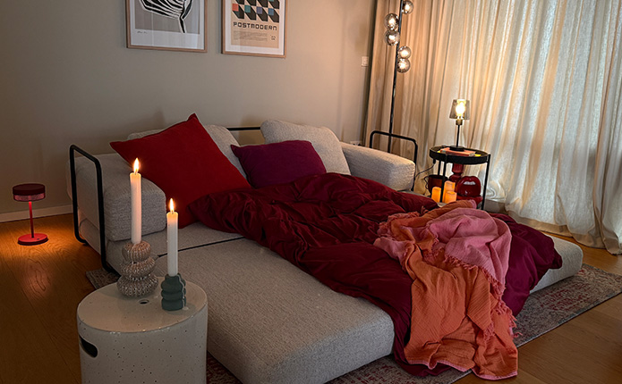Ausgeklapptes Sofa mit dunkelroter Bettwäsche, Kissen, Kuscheldecke und Lichtern