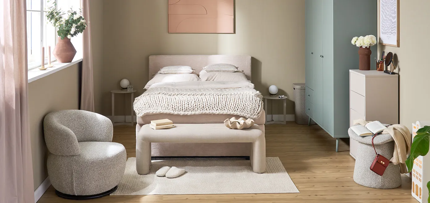 Schlafzimmer mit Polsterbett, abgerundeter Posterbank und Drehsessel in Beige, pastellakzente in Kleiderschrank und Deko