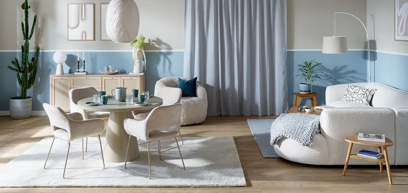 Wohnzimmer mit weißem Ecksofa mit runden Formen und Boucléstoff, Wandfarbe horizontal aufgeteilt in der Mitte, unterer Teil in Hellblau, oberer Teil in Hellgraubeige, runder Esstisch mit vier hellgrauen Polsterstühlen, weißer Teppich, Kommode aus hellem Holz und Rattan