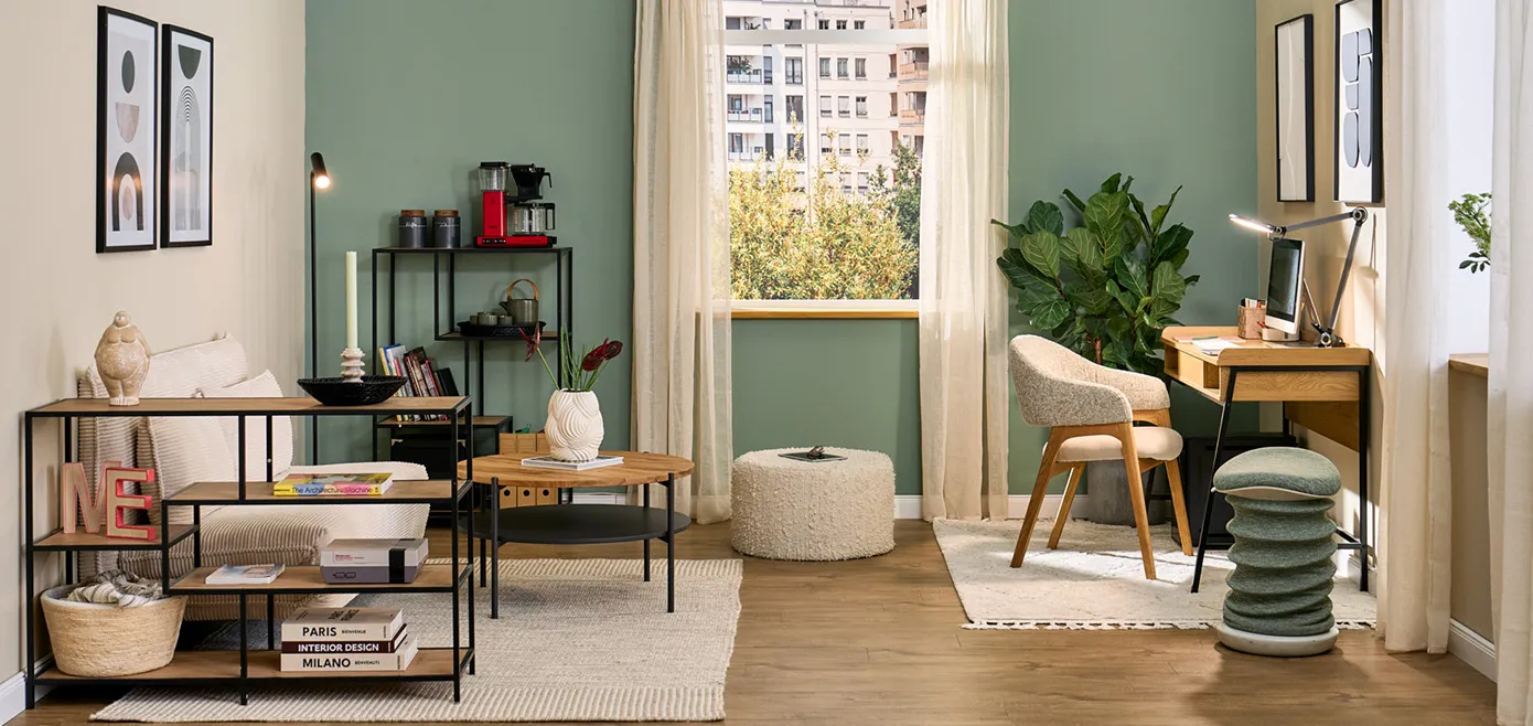Kleines Zimmer mit Schreibtisch aus Holz und schwarzem Gestell, Polsterstuhl in beige, grüner Hocker, auf der anderen Seite beige kleine Schlafcouch mit rundem Couchtisch und zwei Standregalen