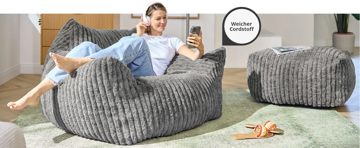 Frau ist sitzt auf Boneless-Sessel aus grauem Cord. Daneben passender Cord-Hocker in Grau in einem Wohnzimmer. Hinweis: Weicher Cordstoff.