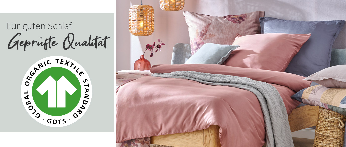 Hinweistext: Für guten Schlaf, geprüfte Qualität, Logo GOTS: Global Organic Textile Standard. Schlafzimmer mit Holzbett mit Bettwäsche in Rosé, Blau und Rosa mit Blumenmuster
