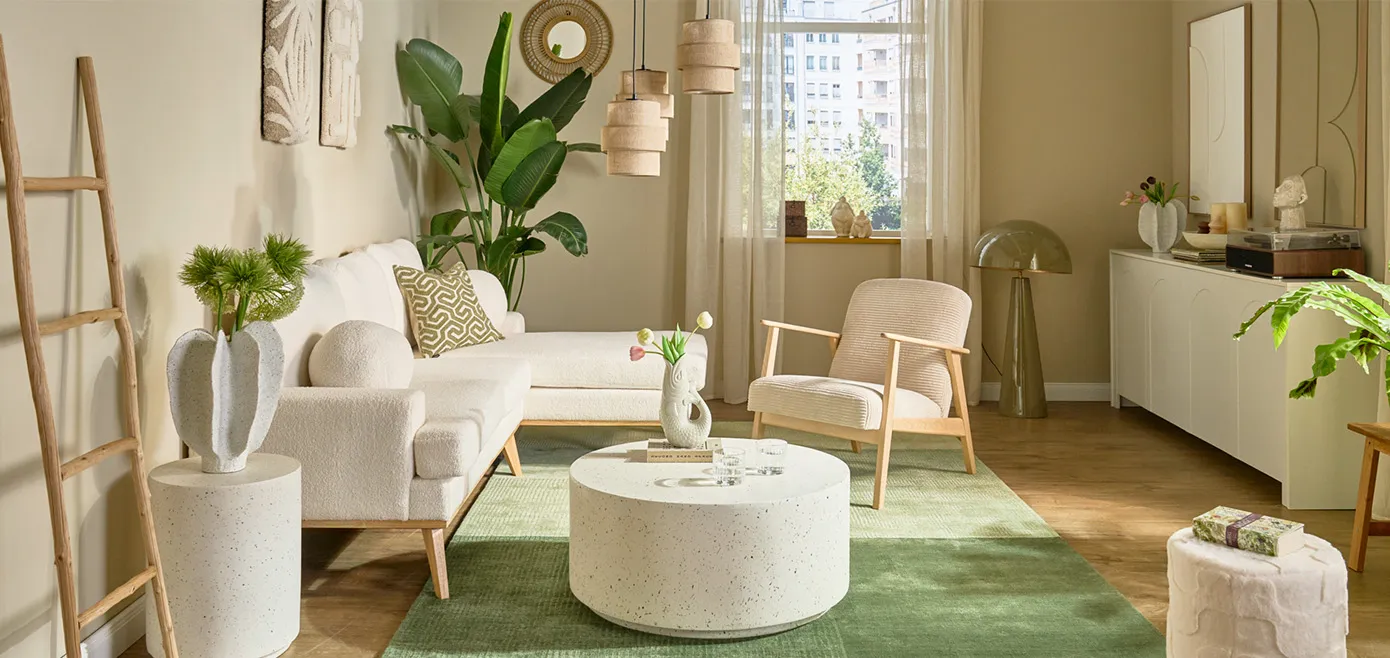 Weißes Bouclé-Ecksofa mit Holzfüßen, grüner Teppich, runder weißer Couchtisch in Steinoptik mit passendem rundem Beistelltisch, beiger Cord-Sessel mit Holzgestell, beige Kommode Pendelleuchte aus Leinen, große Pflanze in der Ecke
