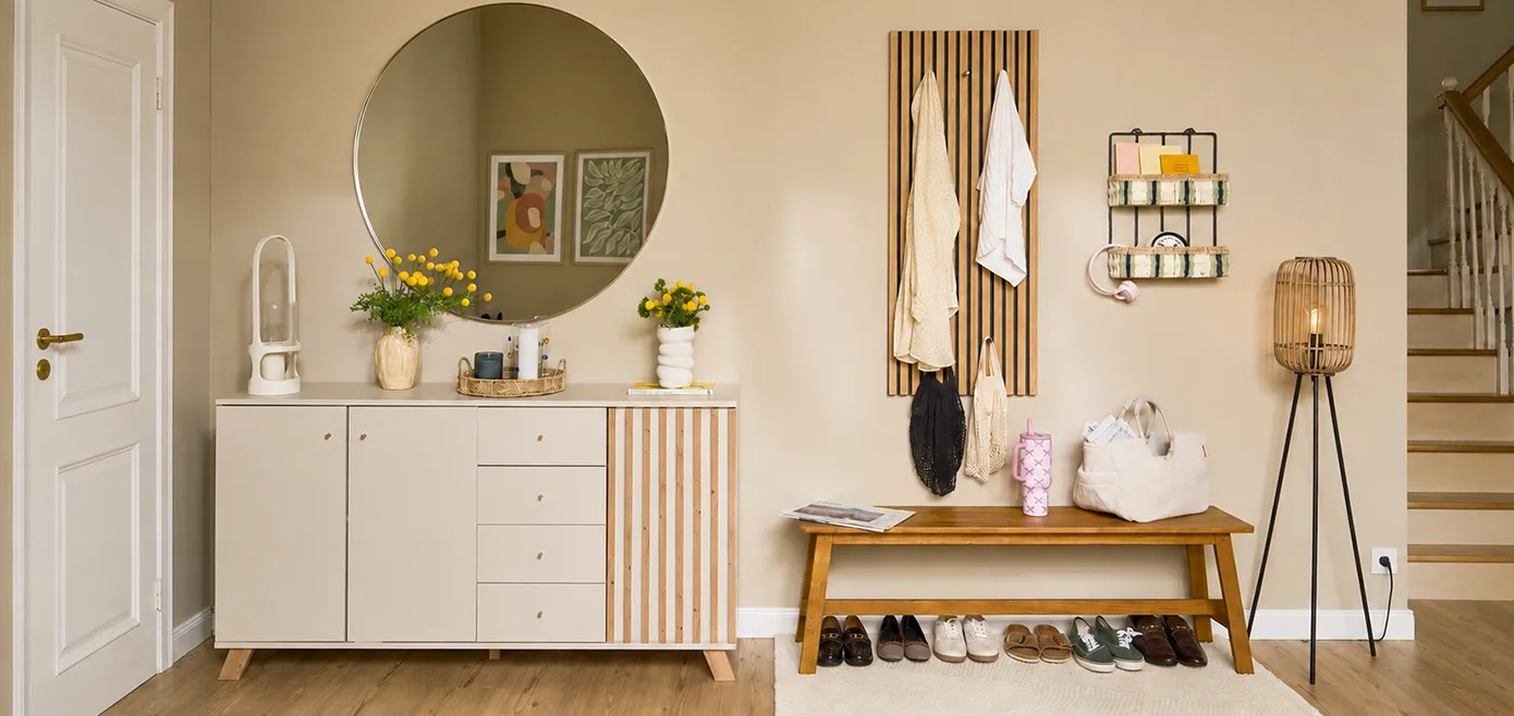 Lange Wand im Flur mit Treppe nach oben, vor cremefarbener Wand cremefarbene Kommode mit Holzlamellen-Applikationen, darüber großer runder Wandspiegel, neben Kommode Holzbank mit Schuhen darunter, Wandgaderobe aus Holz ebenfalls in Lamellenoptik, Stehleuchte und kleine Wandregal