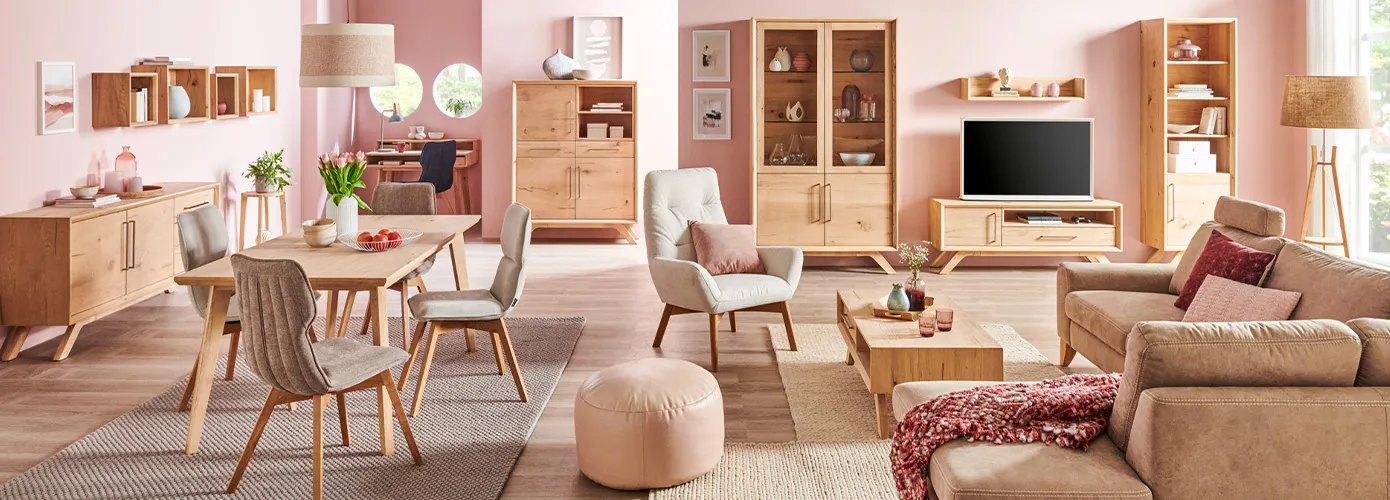 Wohnzimmer im Skandi-Stil mit Holzmöbeln, bestehend aus Sideboard, Wandboard, Esstisch mit grauen Polsterstühlen mit Holzgestell, cremefarbigen Sessel mit Holzgestell, Couchtishc beigem Ecksofa mit Kissen und Plaid, beigen Pouf, Schreibtisch, Highboard, Vitrine, Lowboard, Wandregal, Regal, Textilien, Teppiche, Deko.