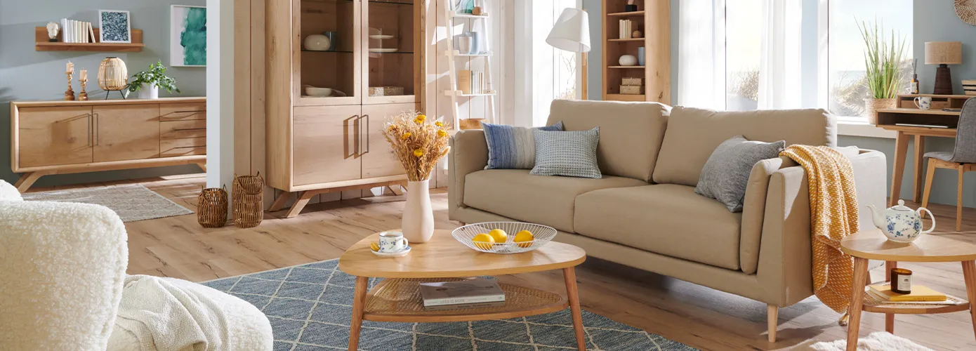 Wohnzimmer mit Möbeln im Skandistil, bestehend aus weißem Sessel, Holz-Sideboard, Holz-Wandboard, Holz-Vitrine, weißem Regal, beigem Sofa mit Kissen, Holz-Couchtisch, Holz-Regal, Holz-Sekretär, Holz-Beistelltisch, Deko