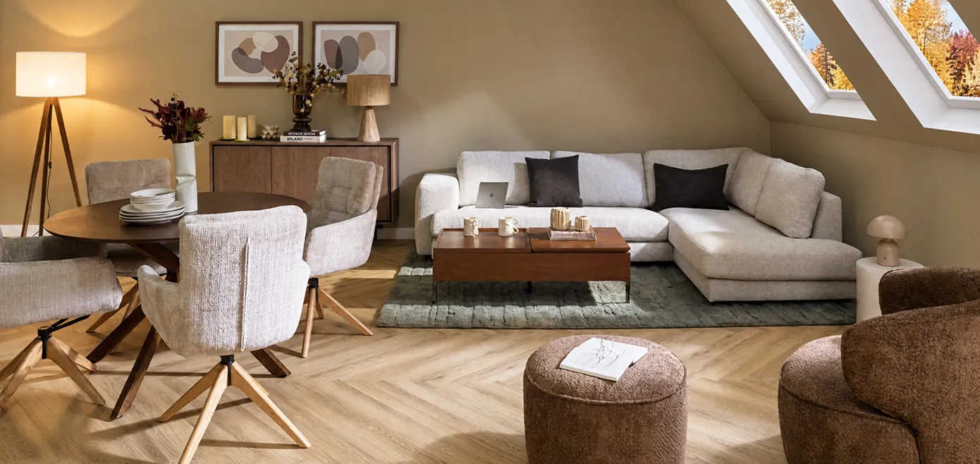 Beige meliertes Ecksofa unter Dachschräge, grüngrauer Teppich, rechteckiger schlichter Couchtisch aus dunklem Holz, runder dunkelbrauner Esstisch mit vier hellgrauen Polsterstühlen, dunkelbraune Kommode