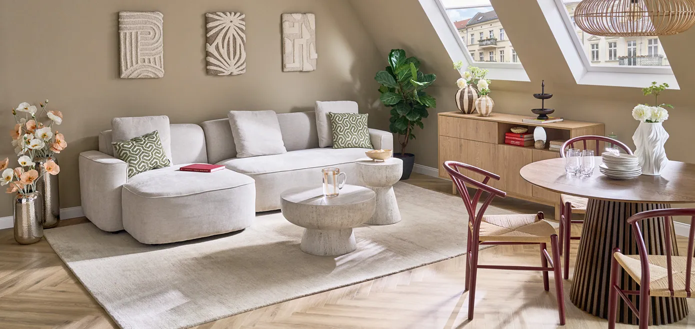 Beiges Ecksofa mit runden Ecken, großer weißer Teppich, runder Couchtisch und passender Beistelltisch in Steinoptik, Stoff-Leinwandbilder an Wand mit verschiedenen Mustern, dunkelbrauner runder Esstisch mit rotbraunen filigranen Stühlen, Kommode aus hellem Holz mit Dekovasen in Braun, zwei große Chrom-Bodenvasen mit Pflanzen