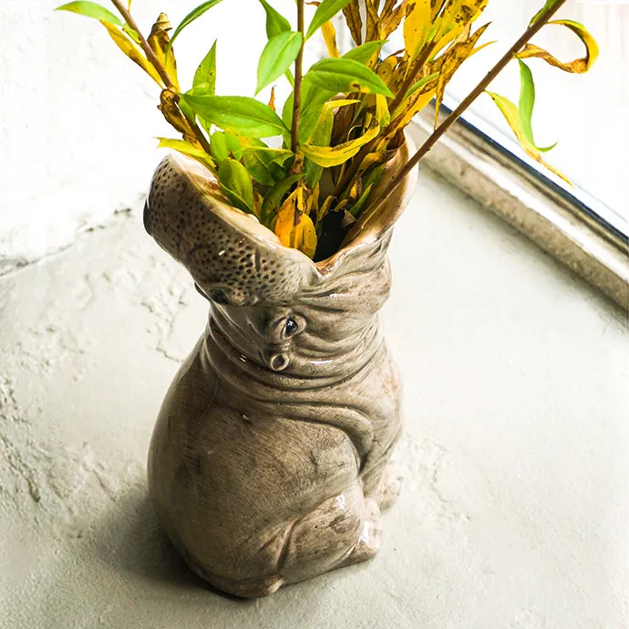 graue Vase in Nilpferdoptik mit grünen Zweigen darin vor Fenster