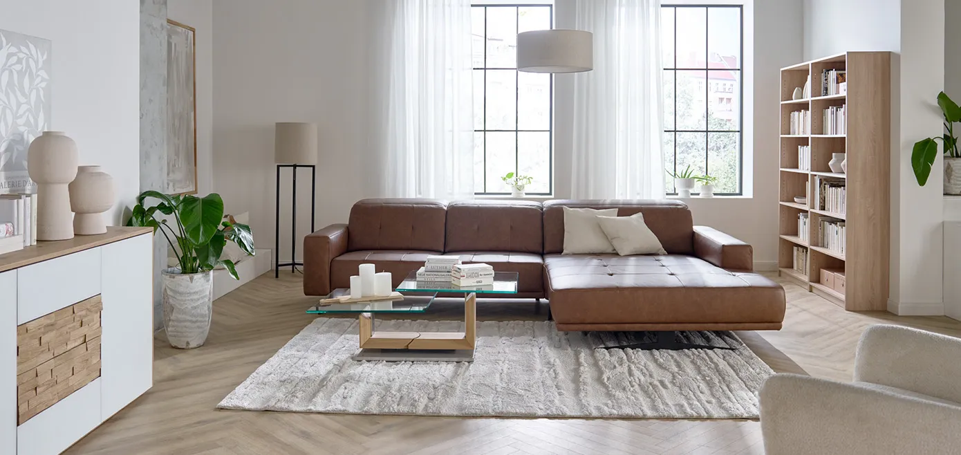 Gradliniges braunes Leder-Ecksofa, flauschiger beiger Teppich, Couchtisch mit dickem Holzfuß und zwei Glasplatten, hohes Holzregal mit Büchern, weißes Sideboard mit Holzschubladen in der Mitte, Steinvasen
