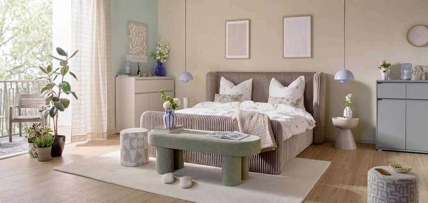 Graubraunes Doppelbett aus Cord mittig vom Raum vor beiger Wand, rechts und links davon hellblaue Pendelleuchten, ovale khakigrüne Sitzbank, runder Pouf in Beige und geometrischen Mustern, zwei Kommoden in salbei und kaschmirfarben, beiger Teppich unter Bett und Sitzbank, verschiedene Pflanzen verteilt in beigen und blauen Vasen