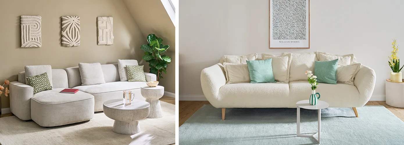 Beiges Ecksofa mit abgerundeten Ecken, grün-beige gemusterte Dekokissen, runder Couchtisch und Beistelltisch in Steinoptik, beige Wand und drei Leinwandbilder in beige und geometrischen Mustern