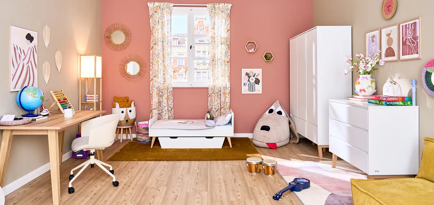 Kinderzimmer mit weißer Kommode, Kleiderschrank und Kinderbett von pinio, Holz-Schreibtisch mit weißem Drehstuhl, Dekobilder an Wand, Hund-Sitzsack und bunte Aufbewahrungskörbe