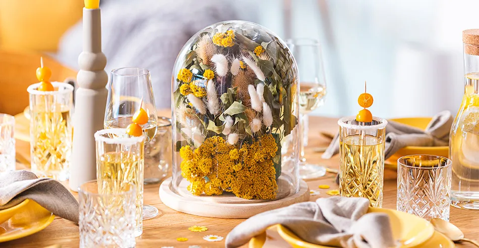 Gedeckter Holztisch mit gelben Tellern, Schüsseln und Stabkerzen, dazu Blumenglocke mit gelben Blumen