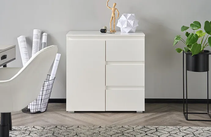 Weiße Kommode mit Deko, weißer Stuhl, schwarzer Drahtkorb mit Papierrollen, schwarze Blumensäule mit Grünpflanze, grauer Teppich mit schwarzem Muster, Deko
