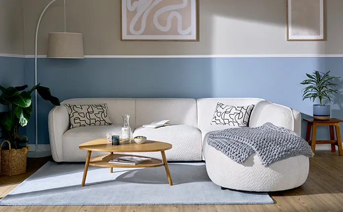 Hälfte der Wand in Blau horizontal, rundes, weißes Ecksofa mit Muster-Dekokissen und graublauem Strickplaid, ovaler Couchtisch aus Holz mit Rattanfläche, hellblauer Teppich