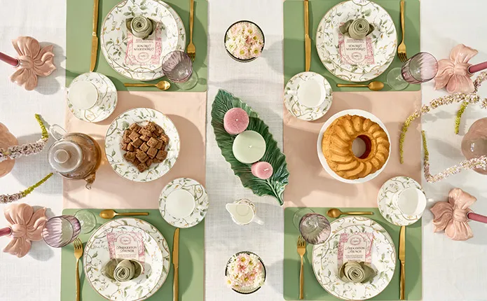 Top-Ansicht: Langer Tisch mit weißer Tischdecke, cremefarbenen Essstühlen und Holzgestell, runde Vasen in Rosa mit Blüten und Pflanzen in weiß und rosa, florales Geschirr in weiß mit grünen Ranken, rosa Schleifen Kerzenständern, grüne Eissschalen, Zitronentarte, Kerzen in mint und rosa, mintfarbene Platzsets