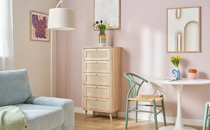 Schmale und höhere Holzkommode aus hellem Holz in Rattanoptik und 5 Schubladen, kleiner runder Esstisch in Weiß, Rattanstuhl mit grünem Gestell, bunte und gestreifte Dekovasen auf Tisch und Kommode