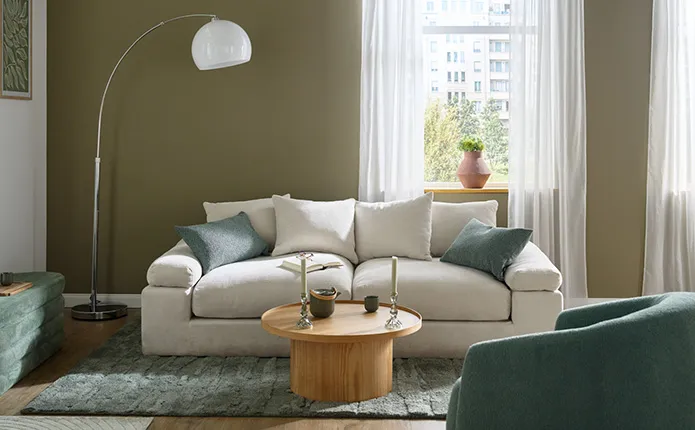 Wohnzimmer mit weißer und olivegrüner Wand, cremefarbenes Sofa als Zweisitzer mit grünen Kissen, daneben eine Polstersitzbank in Grün mit Stauraum, runter Couchtisch aus hellem Holz, grüner Teppich, weiße Bogenleuchte und grüner Sessel aus Boucléstoff