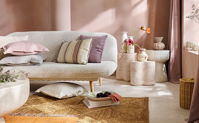 Hellgraues Sofa im Skandistil mit gestreiften und farbigen Kissen in Beige, Rosa und Lilatönen, geflochtener Teppich und Aufbewahrungskorb, verschiedene Beistelltische in Steinoptik mit verspielten Dekovasen