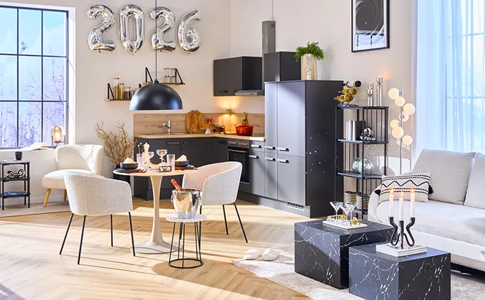Eck-Einbauküche in Dunkelgrau, dazu kleiner runder Esstisch in Kaschmirfarben und grauen Stoffstühlen, kleiner Beistelltisch mit Sektkühler und gedecktem Tisch, Marmor-Couchtische in Schwarz, Diskokugeln und Chromdeko, silberne 2026 Luftballons an der Wand