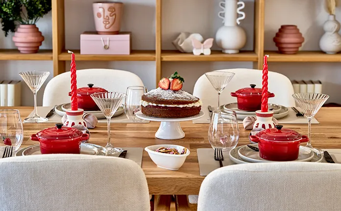 Holztisch mit beigen Stühlen, Tischdeko mit roten und beigen Farbtönen mit Mini-Kochtöpfen in Rot, beiges Keramikgeschirr, Martinigläser, Vase im Boho-Stil, Erdbeerkuchen, Servietten als Herz gefaltet, rote Stabkerzen