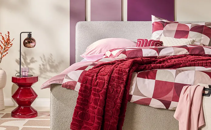 Hellgraues Bett mit gemusterter Bettwäsche in Beerentönen, roter Glas-Beistelltisch