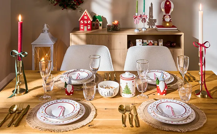 Gedeckter Holztisch mit rot-weißem Weihnachtsgeschirr, goldenem Besteck, runden Platzdeckchen, Weingläsern mit Goldrand, goldene Kerzenständer mit roter und grüner Schleife und Stabkerzen in Rot