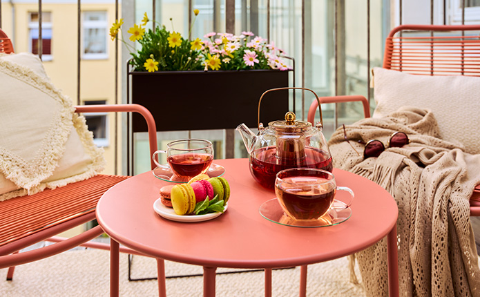 Detailansicht kleiner Tisch mit Teekanne, Teetassen und Macarons. Im Hintergrund Pflanzenkübel
