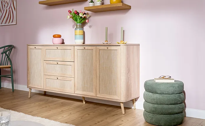 Breites Sideboard aus hellem Holz in Rattanoptik mit drei Schubladen und drei Türen, darauf bunte und gestreifte Dekovasen und Blumenkerzenständer mit grünen Stabkerzen, neben Sideboard ein runder grüner Pouf