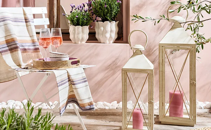 Zwei weiße Laternen mit pinken Stumpenkerzen, weißer Klappstuhl und gestreifter Decke und Serviertablett mit Aperolgläsern