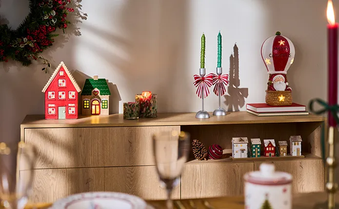 Holz-Sideboard mit weihnachtlicher Deko wie ein rotes Haus, ein beiges Haus mit grünem Dach beleuchtet, Schleifen-Kerzenständern und rot-weiße Dekofigur Weihnachtsmann in Heißluftballon