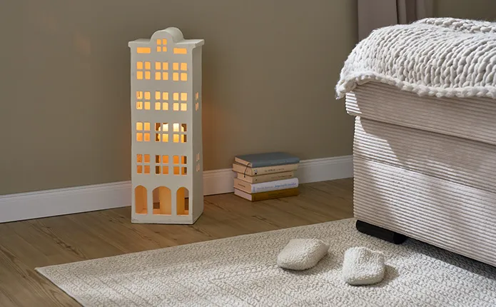 Beiger Teppich mit Hausschuhen, daneben beiges Dekohaus mit großer Kerze zur Beleichtung