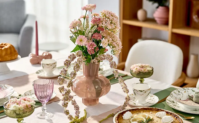 Bauchige Glasvase in Rosa mit Blumen, drumherum der gedeckte Tisch