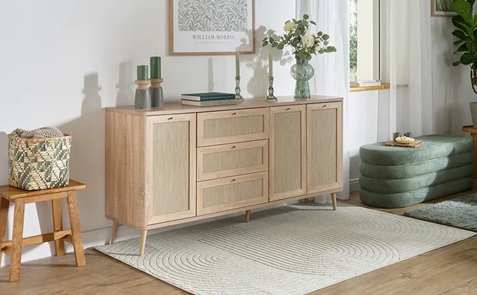 Sideboard in Rattanoptik und hellem Holz, daneben Hocker aus Holz mit gemusterten Aufbewahrungskorb, Struktur-Teppich in leichtem Grünton, Sitzbank in Khakigrün, Vasen und Kerzenständer in Grüntönen