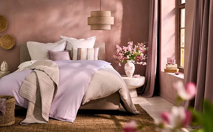 Schlafzimmer mit altrosa Wänden, Pendelleuchte aus Leinen, Polsterbett mit lavendel und beigefarbener Bettwäsche und gestreiftem Kissen, Beistelltisch in Steinoptik mit Blumenvase