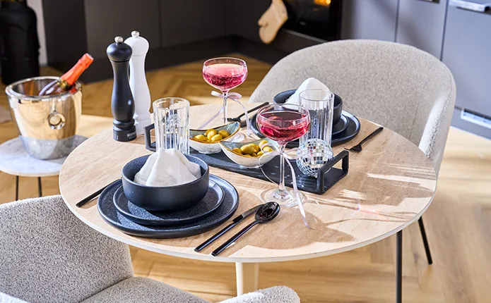 Detailansicht gedeckter, runder Esstisch mit heller Holzplatte, graue Stühle, dunkelblaues und schwarzes Geschirr und Besteck, schwarze Servierplatte mit Schälchen und Fingerfood und eine Diskokugel, Coupettegläser mit Rotwein und Schleifenband-Verzierung, Beistelltisch mit Sektkühler