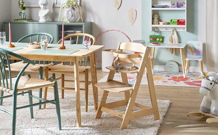 Wohnzimmer im Skandistil mit Holz-Esstisch und passenden Holzstühlen, Hochstuhl, Schaukelpferd, grüngraue Kommode, Spielecke mit kleinem Tisch und Stuhlset mit Pepper Pig, kleiner Teppich mit Blumen, Wandregal mit Spielzeug, farblicher Rundbogen an Wand zur Abgrenzung