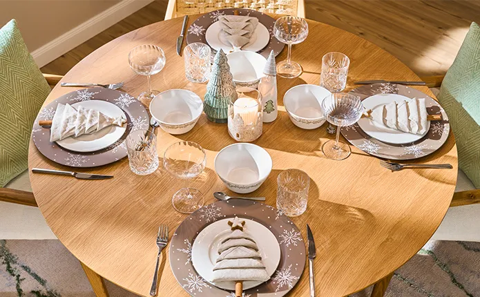 Gedeckter, runder Holztisch mit braunen Dekotellern und weißen Schneeflocken, Servietten als Tannenbaum gefaltet auf weißen Speisetellern, Coupette-Gläser, Schüsseln mit salbeifarbenen Dekozweigen, Deko-Tannenbäume in Eukalyptus-Grün, Beiges Windlicht