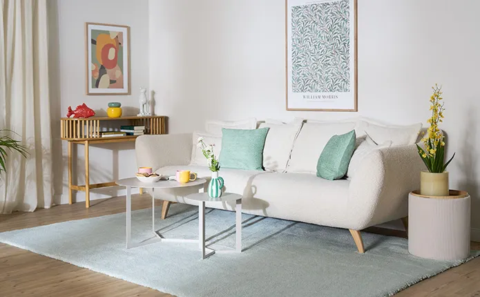 Hellblauer Teppich, Sofa in Weiß und Boucléstoff mit mintgrünen Dekokissen, hellgraue runde Couchtische, Konsolentisch aus Holz, Beistellhocker mit Stauraum