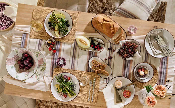 Holztisch gedeckt mit gestreiftem Tischläufer, geflochtenen Platzsets, beiges Geschirr mit schwarzem Rand und Geschirr mit rosa Blumenmuster, Schälchen mit Fingerfood und Spargel, kleine Teelichthalter in Blütenform