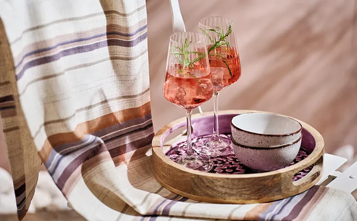 Detailansicht rundes Serviertablett mit lila Blumenmuster, zwei Aperolgläser und Schälchen auf weißem Garten-Klappstuhl und gestreifter Decke