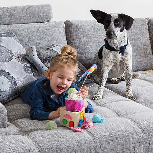 Mädchen mit Eisbecher und Hund auf Sofa, Eiskugeln liegen auf dem Polster
