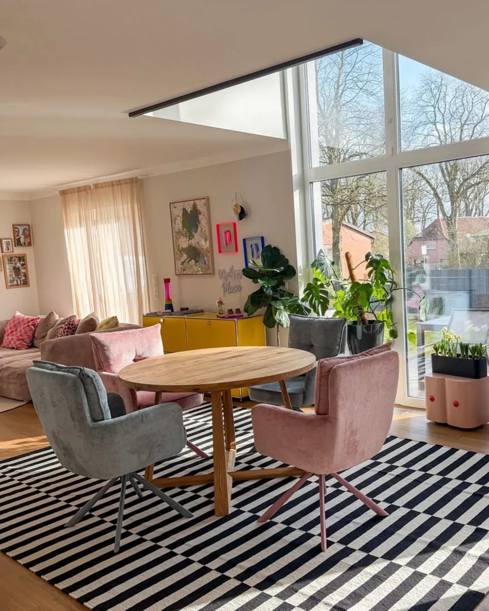 Runde rosa Glasvase mit Frühlingsblumen auf Holz-Couchtisch, dahinter grau-beiges Sofa mit beigen Kissen und Rautenmuster