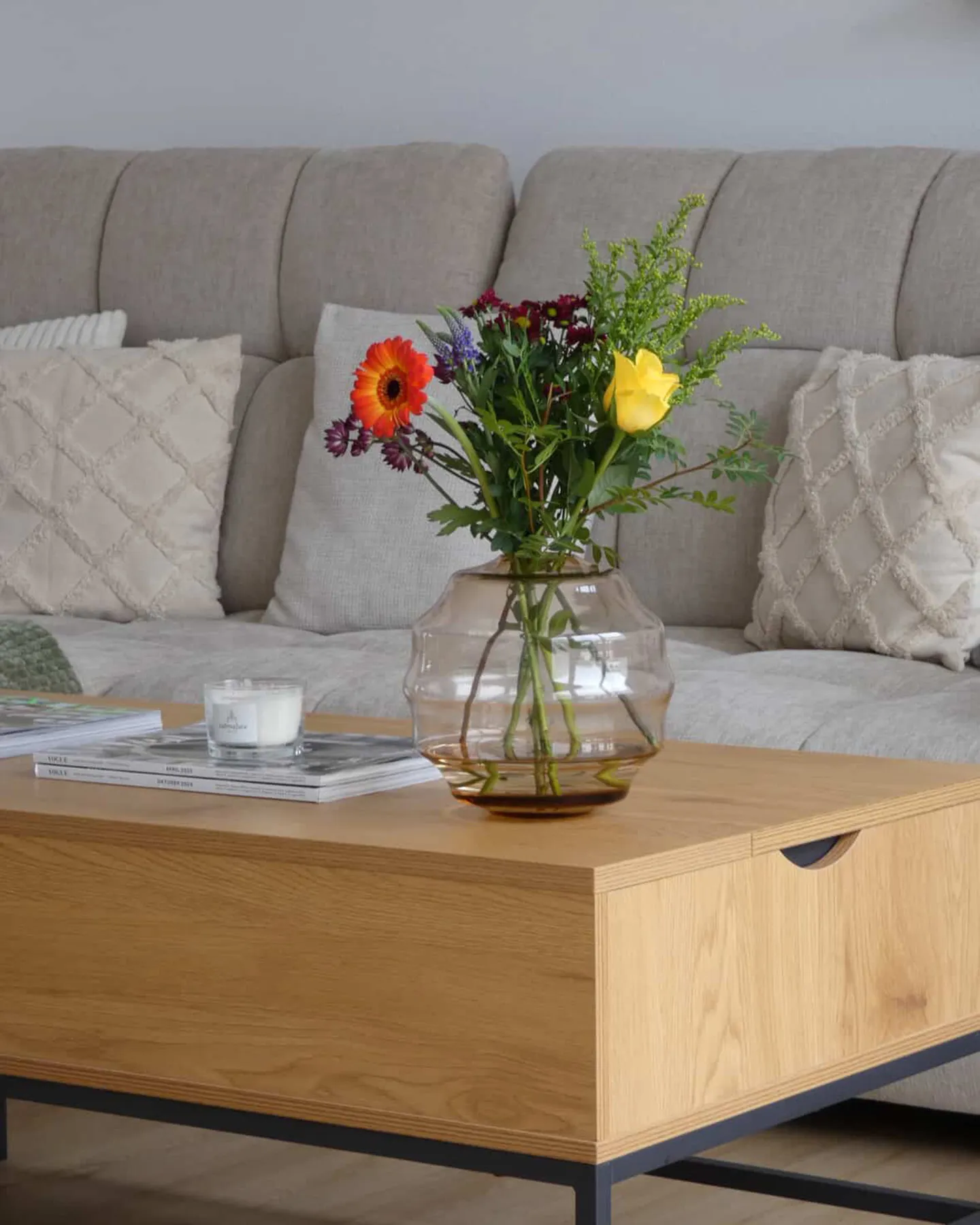 Runde rosa Glasvase mit Frühlingsblumen auf Holz-Couchtisch, dahinter grau-beiges Sofa mit beigen Kissen und Rautenmuster