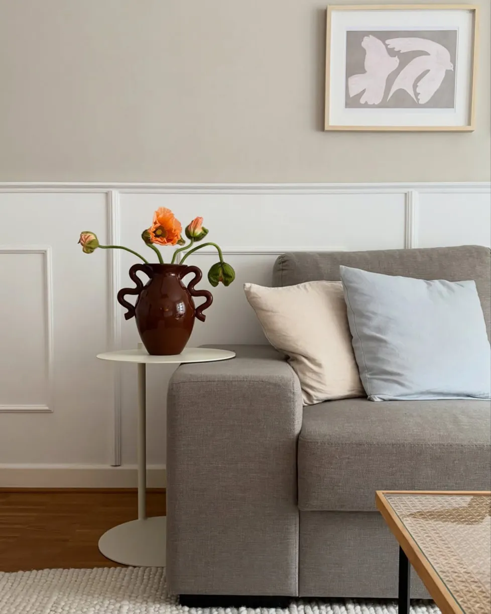 Beige Wand mit horizontaler weißer Wandverkleidung mit Lamellen, davor graues Sofa, kleiner runder beiger Beistelltisch mit braunroter Vase und geschwungenen Henkeln mit orangenen Blumen, Dekokissen in Hellblau und Creme