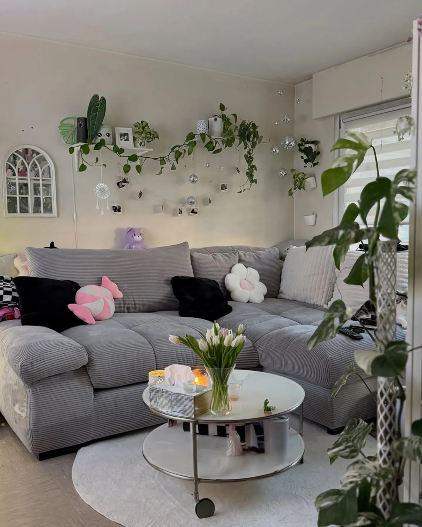 Hellgraues Ecksofa aus Cord mit weißem runden Teppich, runder kleiner Couchtisch auf Rollen in weiß mit Chromgestell, schwarze, weiße Dekokissen, ein Dekokissen in Bonbonform und eins in Blumenform in rosa-weiß, viele Pflanzen und Tulpen, Diskokugeln an Wand und Wandregalen