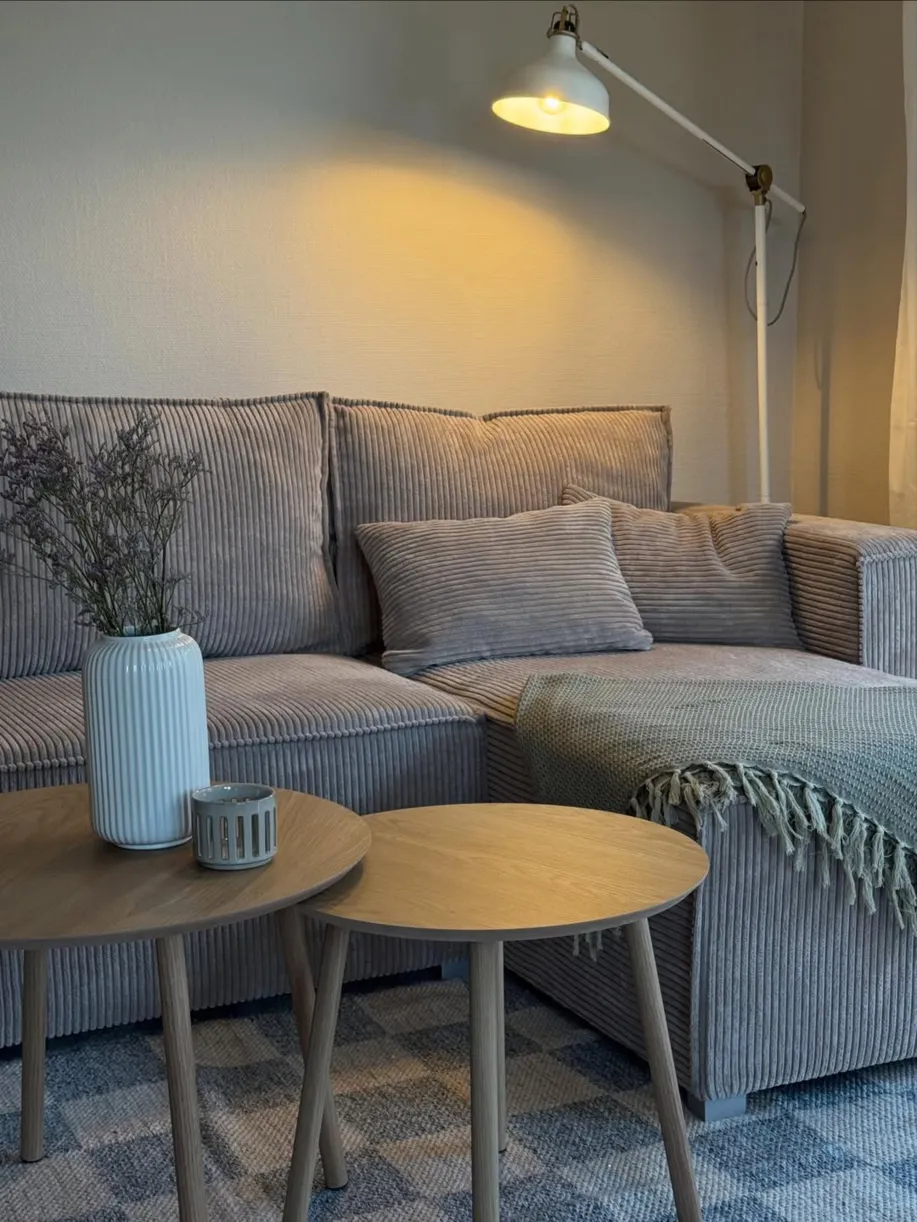 Hellgraues Ecksofa aus Cord mit dunklem Holzboden und schwarzen Accessoires wie Regal und Pendelleuchte