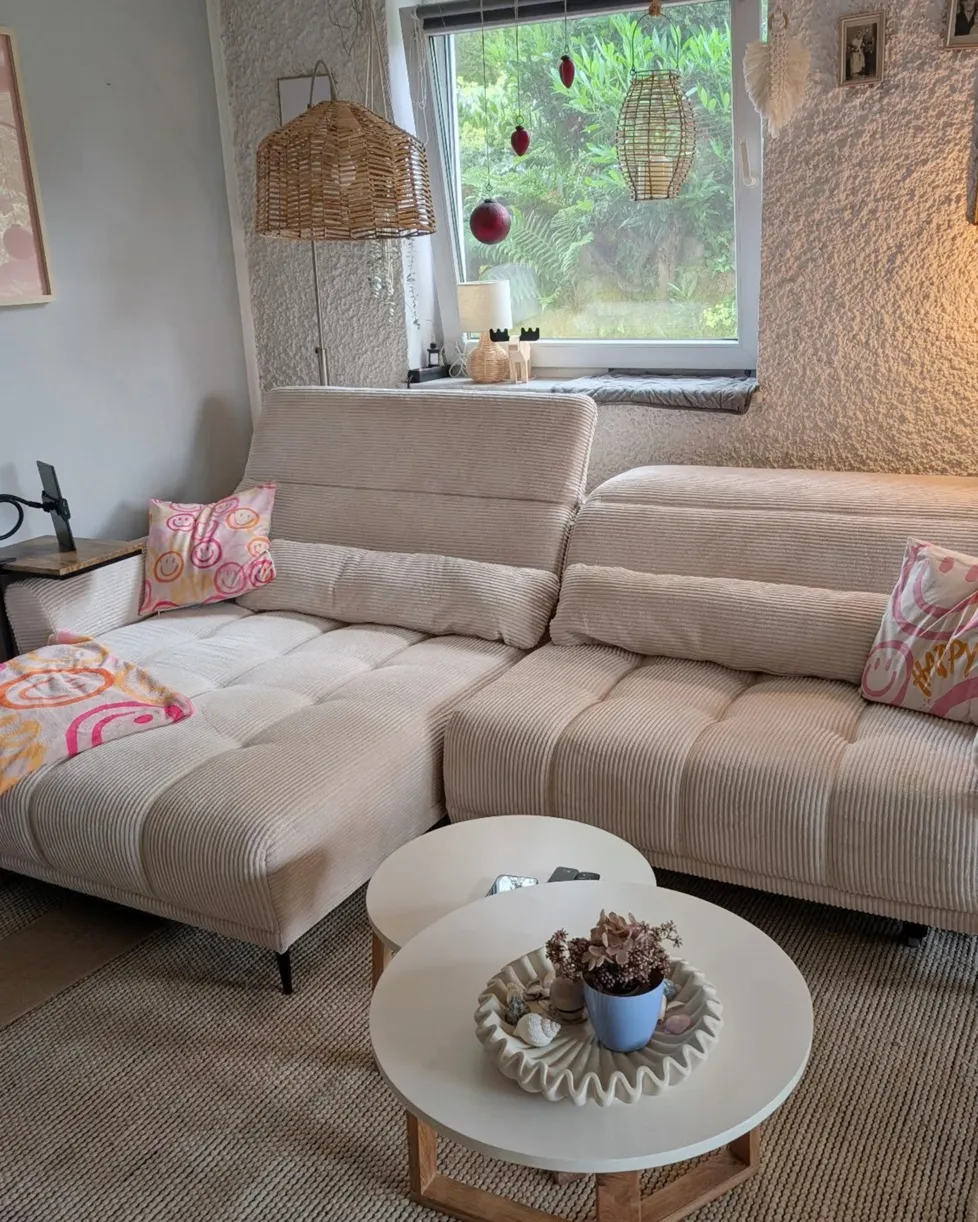 Runde rosa Glasvase mit Frühlingsblumen auf Holz-Couchtisch, dahinter grau-beiges Sofa mit beigen Kissen und Rautenmuster