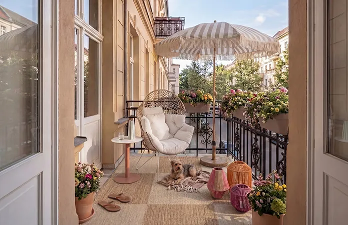 Balkon mit schwarzem Geländer, beigefarbenem Teppich, Beistelltisch rund mit rostfarbenem Bein und beiger Platte, Loungesessel mit beiger Polsterung und weißen dünnen Beinen, bunten Windlichtern in Rosa und Orange, gestreifter Sonnenschirm mit Fransen
